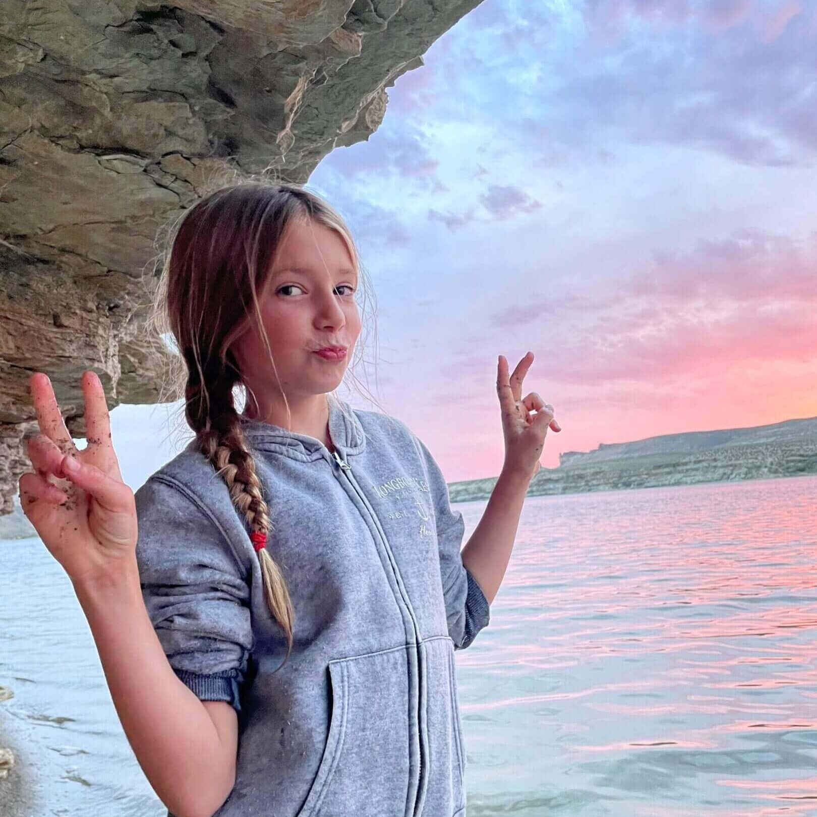 Girl with braided hair making peace signs by a lake at sunset.